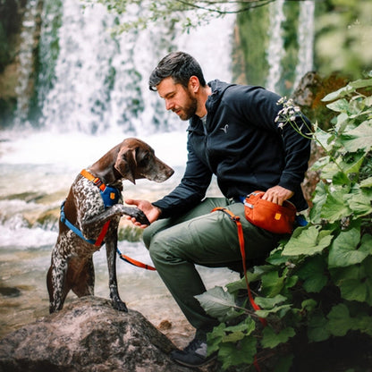 Hund gibt Pfote und Mensch mit Alpha 360 Hüfttasche in Terrakotta sucht nach Leckerlis bei einer Pause in der Natur.